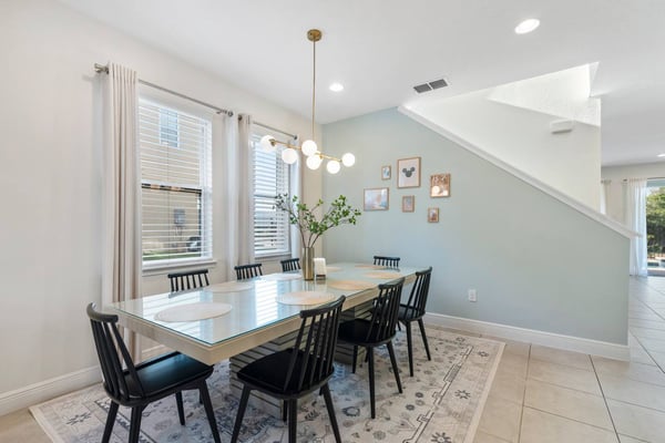 Bright dining room with white table, black chairs, pendant light, pale blue accent wall, framed artwork, and staircase to the right.