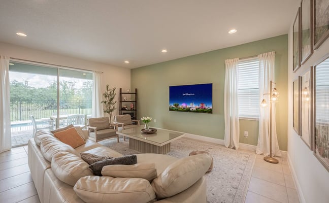 Open-plan kitchen and living room with tile flooring, white cabinetry, island seating, and glass doors opening to pool and lawn area.